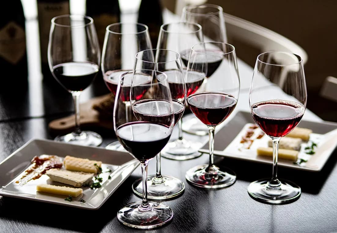 Flera glas med rödvin uppställda på ett bord tillsammans med tallrikar fyllda med olika ostar, redo för vin- och ostprovning.